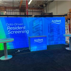RENTAL: RE-2111 Inline Design with Single-Sided Lightbox with Flat Canopies with Black Pillowcase Fabric Covers, (2) RE-704 Charging Station Tables, RE-1567 Backlit Reception Counter, SEG Backlit Fabric Graphic, and Vinyl Applied Graphics