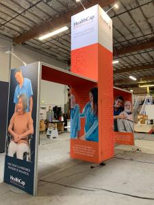 RENTAL: Modified RE-9195 Island Design with Gravitee Storage Closet @ 42" x 42" x 190" H with Locking Door, (2) Gravitee Arch Canopy Structures @ 84" Long x 95" High, (1) Large Monitor Mount, 43" Monitor, 30" W x 16" D White Laminated Shelf, and SEG Fabri