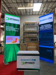 RENTAL: Modified RE-9129 Island Design with (2) 4' Wide x 12' High Double-Sided Lightboxes with Boomerang Baseplates, (2) Large Monitor Mounts, (2) 50" Monitors with HDMI Cables, RE-1576 White Laminated Reception Counter at 48" Wide x 24" Deep x 38" High