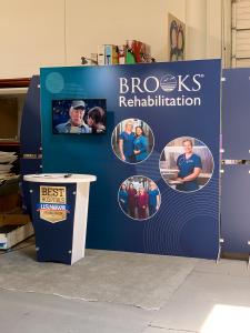 RENTAL: RE-2139 Design with 220" Wide x 90" High Backwall, (2) Monitor Mounts, (4) LED Arm Lights, RE-1201 Tapered Reception Counter, SEG Fabric Graphic, Direct Print Sintra Accent Wing Graphics, and Direct Print Sintra Reception Counter Graphic -- View 3